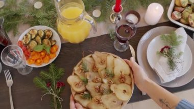 Lezzetli yemekler, taze portakal suyu ve kızılcık suyuyla dolu ve çam dalları ve mumlarla süslenmiş Noel masasının üzerine fırında armut ve ceviz koyan kadın eli tabağı.