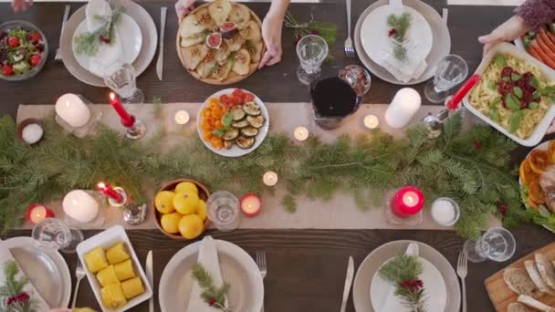 クリスマスディナーの準備家族のトップビュー 火入れのろうそくと松の枝から作られた中心部で飾られたテーブルの上においしい料理を置いています — ストック動画