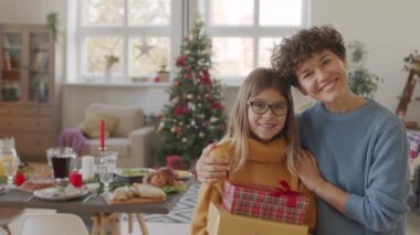 Kısa kıvırcık saçlı ve Noel hediyeleriyle birlikte oturma odasında poz veren neşeli bir genç kadının portresi. Yemek dolu yemek masası ve arka planda Noel ağacı
