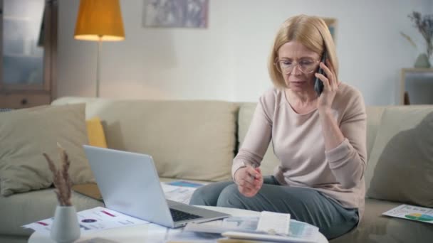 金融上の問題を抱えながら電話で話しながら財務諸表や書類をめがねの中の心配な中高齢原因女性の映像の並び — ストック動画