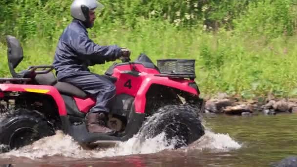 ゆっくり追跡のひげを生やした男でヘルメットに乗って赤いクワッドバイク川を通って森の中 — ストック動画
