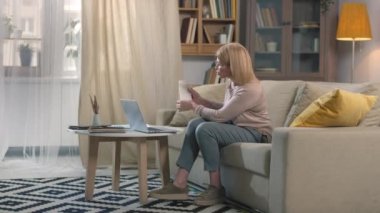 Orta yaşlı beyaz bir kadının iş ortağıyla bilgisayarında video görüşmesi yaparken faturaları ve ödemeleri evde çalışırken göstermesi.