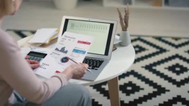 Yazılı çizelge ve şemalara bakarak aylık zorunlu harcamalara göre hesap çizelgesini açmış beyaz bir kadının dizüstü bilgisayarının önünde oturduğu arka plan görüntüleri.