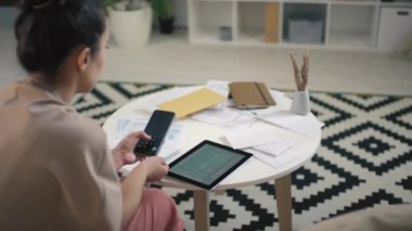 Parlak oturma odasında oturan tabletin mali durumunu analiz ederken telefonunda hesap makinesi uygulaması kullanan melez bir kadının arka plan görüntüsü.