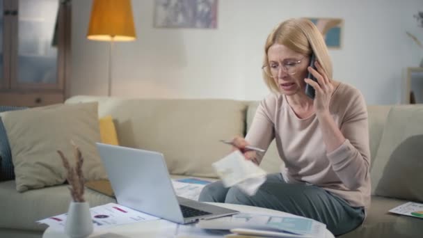 電話で話す成功した中年の白人女性リビングルームの机の上の文で文書の山を通過書類を行う中で財政問題を議論 — ストック動画