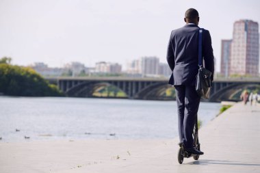 Şehir manzarasına karşı nehir kenarında yürürken, resmi giyinmiş, elektrikli scooterın üzerinde duran genç ve zarif işadamının arka planı.