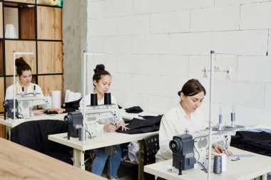 Sıraya dizilmiş genç terziler yeni mevsimlik koleksiyon üzerinde çalışırken masa başında oturup elektrikli dikiş makineleri kullanıyorlar.
