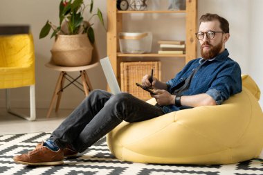 Gözlüklü ve rahat koltukta oturup yeni logolar çizerken sana bakan serbest çalışan genç bir erkek tasarımcı.