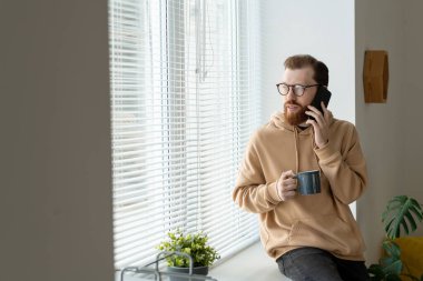 Genç sakallı iş adamı, günlük giysiler ve gözlüklerle akıllı telefondan konuşuyor, pencere kenarında oturuyor ve evde kahve içiyor.