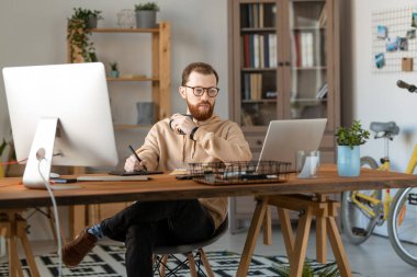 Genç ciddi erkek serbest tasarımcı içki içerken dizüstü bilgisayara bakıyor ve yeni logo çizmek için tablet ve stil kullanıyor.