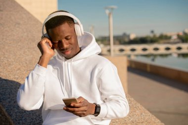 Afrika etnik kökenli mutlu genç sporcu şehir ortamında nehir kenarında uzanıyor ve kulaklıkla müzik dinliyor.