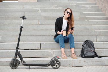 Elinde bir bardak kahveyle merdivenlerde oturmuş akıllı telefondan müzik dinleyen çağdaş bir bayan öğrenci.