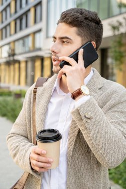 Beyaz gömlekli ve bej ceketli genç zarif bir adam içki içiyor ve müşterisiyle ya da iş arkadaşıyla akıllı telefondan konuşuyor.