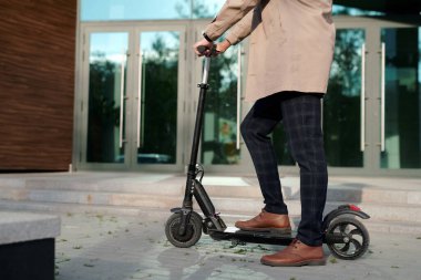 Trençkotlu ve pantalonlu işadamlarının düşük kesimi ofis merkezinin girişinde dikilirken elektrikli scooterın saplarından tutuyorlar.