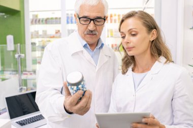 Whitecoat ve gözlüklü kıdemli eczane çalışanı genç sarışın yardımcı hap şişesini gösterirken karakteristik özelliklerini anlatıyor.