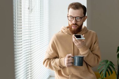 Güncel giysili ve gözlüklü genç adam pencere kenarında oturup içki içerken akıllı telefondan sesli mesaj kaydediyor.