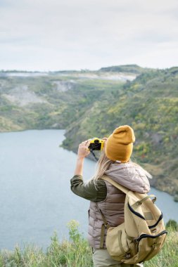 Hafta sonu gezisi sırasında gölün önünde fotoğraf makinesiyle gezen sırt çantalı genç bayanın arka görüntüsü.