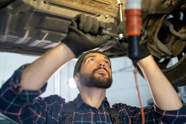 Eldiven ve üniformalı genç sakallı tamirci arabanın altında çalışırken el aletini kullanıyor ve işlem sırasında alt kısmına bakıyor.