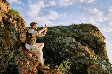 Handsome bearded hiker in vest sitting on cliff rock and photographing landscape on smartphone clipart