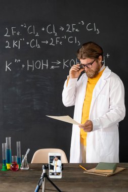 Whitecoat 'taki çağdaş öğretmen, online seyircisi için kimya dersi kaydı yapıyor. Akıllı telefon kamerasıyla tahtaya karşı.