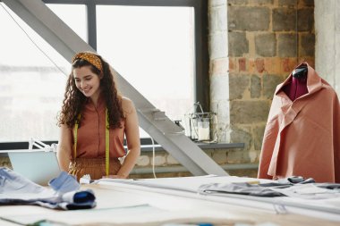 Yeni müşteriler beklerken ve moda koleksiyonu üzerinde çalışırken dizüstü bilgisayara bakan neşeli yaratıcı tasarımcı.