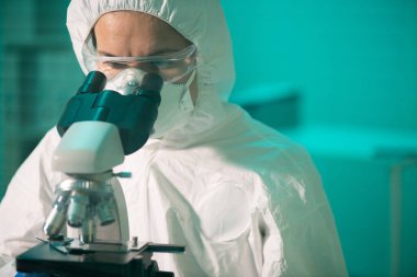 Koruyucu tulumlar, solunum cihazı ve gözlük takan çağdaş araştırmacı yeni kimyasal maddeler üzerinde çalışırken mikroskoba bakıyor.