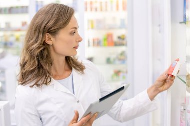 Dijital tabletli genç sarışın eczacı eczanede çalışırken kırmızı kutudaki yeni ilaçlar hakkında bilgi ediniyor.