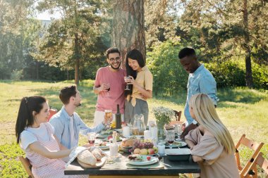 Kültürler arası mutlu genç çift arkadaşlarıyla dışarıda yemek yerken servis edilen masanın önündeki çam ağacının gövdesinde kadeh kaldırıyor.