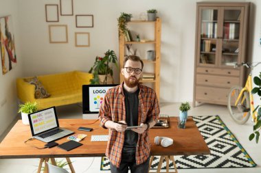 Ciddi yaratıcı tasarımcı kameranın önünde duruyor ve size logolu bilgisayar ve dizüstü bilgisayarın arka planında bakıyor.