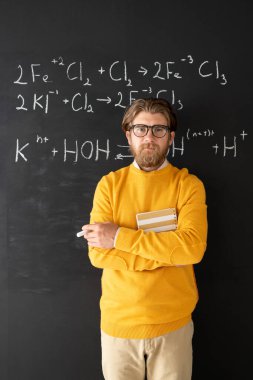 Çevrimiçi ders sırasında akıllı telefon kamerasına bakarken karatahtadaki kimyasal formüle işaret eden günlük kıyafet giyen sakallı kimya öğretmeni.