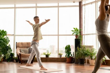 Spor kıyafetli genç bir adam, kollarını açmış yoga egzersizi sırasında öndeki kadına bakıyor.