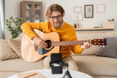 Mutlu sakallı sarışın adam akıllı telefon kamerasına bakarak online izleyicilere müzik yapmayı öğretiyor.