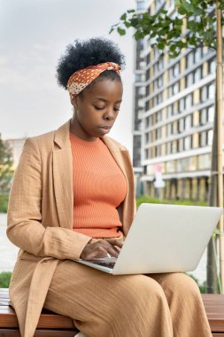 Akıllı gündelik giysiler içinde genç zarif bir iş kadını ofis binalarına karşı parktaki ahşap bankta otururken dizüstü bilgisayarda daktilo kullanıyor.