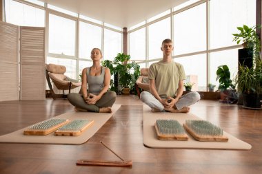 Spor kıyafetli genç bir çift tahta yastıkların önünde nilüfer çiçeği pozu veriyor. Tırnakları ve aromatik özlü çubuklar tütütüyor.