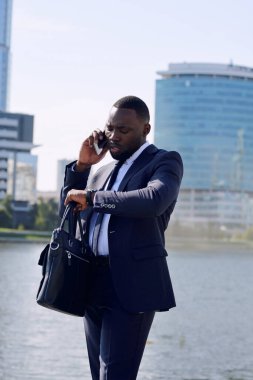 Genç, zarif Afrikalı işadamı veya temsilci, şehir ortamının karşısında dururken, nehir kenarında cep telefonuyla müşterilerine danışıyor.