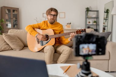 Genç müzik öğretmeni online ders sırasında dinleyicilerine gitar çalarken akıllı telefon kamerasının önünde oturuyor.