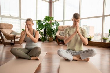 Spor kıyafetli genç bir çift, rahatlama egzersizi sırasında elleri yüzleri tarafından birleştirilmiş nilüfer çiçeği pozunda oturuyorlar.