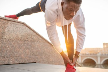 Beyaz kapüşonlu ve siyah şortlu genç aktif Afrikalı sporcu nehir kenarındaki çevrede merdivenlerde egzersiz yapıyor.