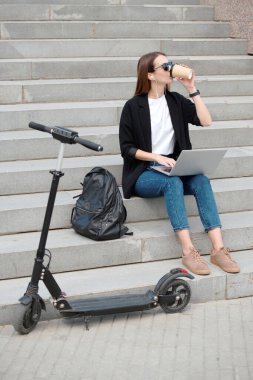 Gündelik kıyafet ve güneş gözlüklü genç bir öğrenci merdivenlerde bir bardak kahve içerken dizüstü bilgisayar kullanıyor ve kulaklıkla müzik dinliyor.