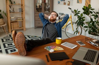 Rahat giyimli ve kulaklıklı genç tasarımcı iş yerinin önünde müzik dinlerken bacaklarını masada tutuyor.