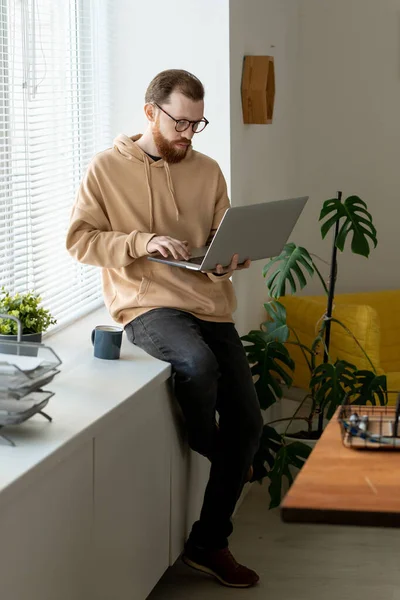 年轻而严肃的自由撰稿人坐在窗台上 一边喝咖啡 一边在家里做远程工作 一边用笔记本电脑 — 图库照片
