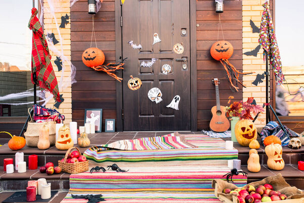 Door of country house decorated with halloween symbols in front of staircase with jack-o-lanterns, spiders, bats, apples and candles