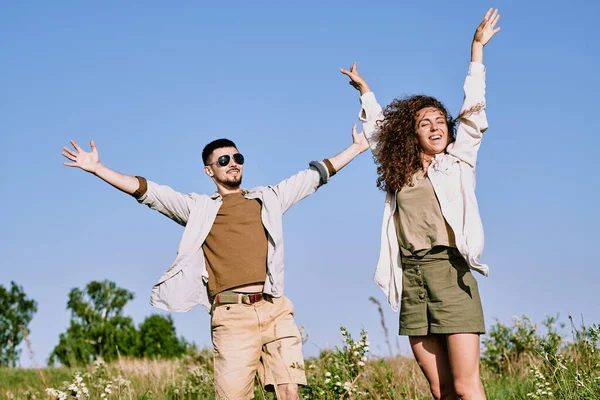 快乐而兴奋的年轻夫妇穿着舒适的衣服 高举双臂站在那里享受乡村旅行 — 图库照片