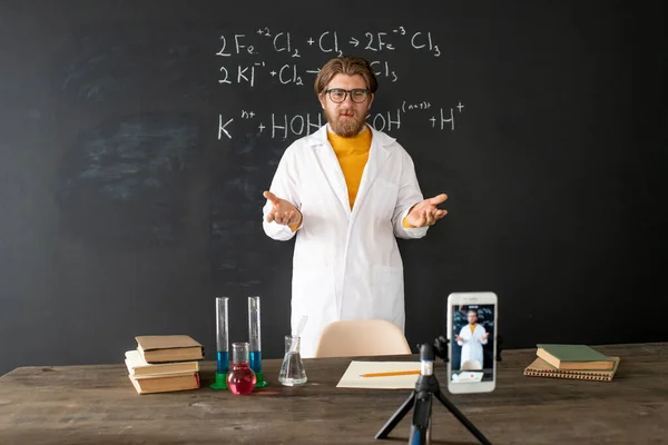 Chemistry teacher in whitecoat standing by blackboard in front of smartphone while shooting himself during online lesson