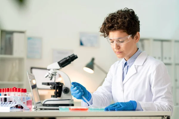 戴着手套的年轻女性实验室工作者 坐在笔记本电脑前的桌子旁 拿着三种物质的样品 — 图库照片