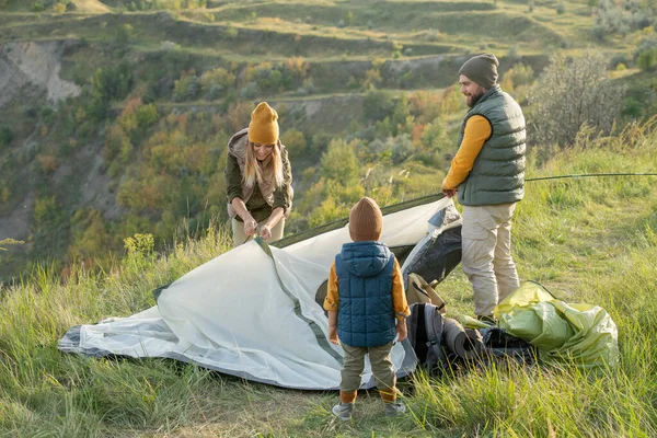 这对年轻夫妇和他们的小儿子站在山顶上的绿草上 把帐篷放在摄像机前的地面上 — 图库照片