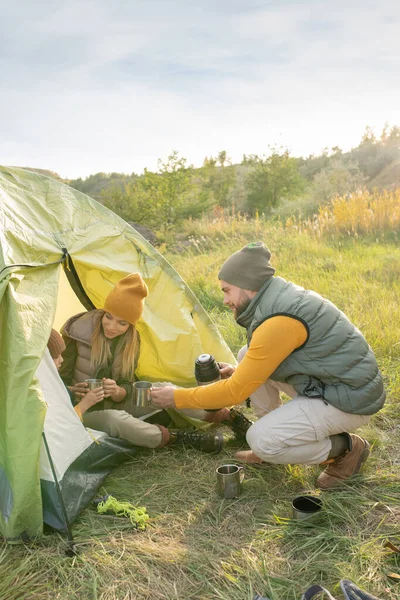年轻人一边蹲在帐篷边 一边在自然环境中的草地上吃饭 一边给他的小儿子一杯热茶 — 图库照片