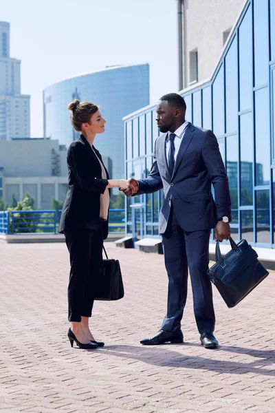 Young successful intercultural business partners in formalwear shaking hands while standing on sidewalk against modern buildings
