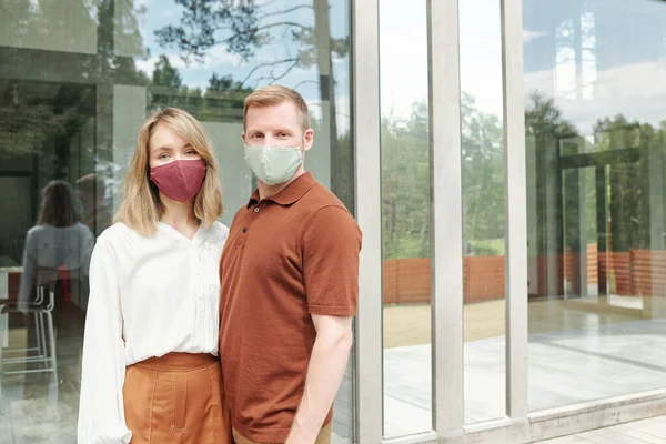 Beautiful couple in cloth masks standing against cottage and embracing while posing for photo