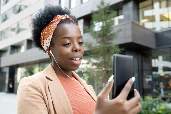在城市通信环境中 快乐的年轻非洲女商人带着耳机看着智能手机屏幕 — 图库照片